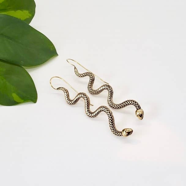 Brass snake-shaped earrings on a white background with green leaves.- Shadowplay Collection Gift Shop