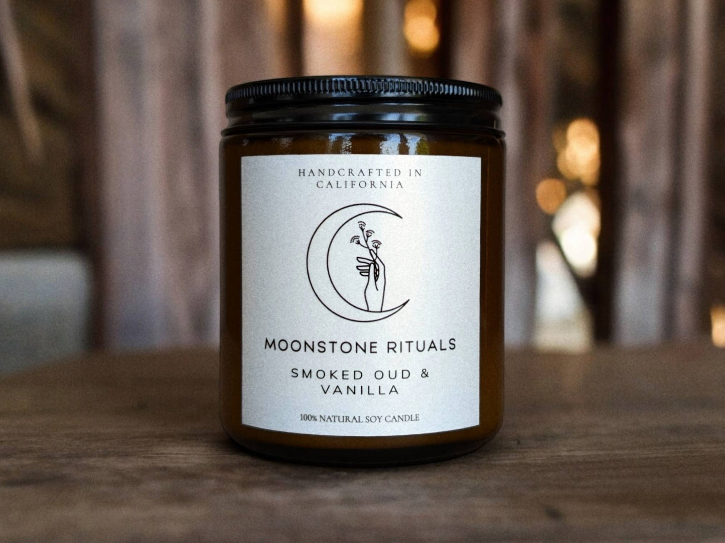 Candle with a label on a wooden surface, featuring 'Moonstone Rituals' and 'Smoked Oud & Vanilla'.